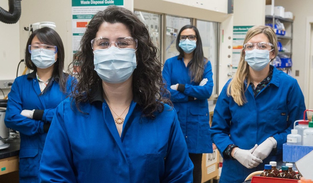 Four women scientists wear marks and glasses in a lab